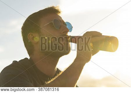A Young Man, With Glasses And A Beard, Drinks Beer From A Bottle, While Enjoying The Landscape And T