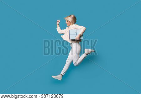 Caucasian Woman Running With A Present On A Blue Studio Wall Wearing White Clothes And Blonde Hair