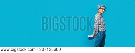 Fashionable Woman In Blue Clothes And Hat Is Posing Near Free Space Holding A Present On A Blue Bann