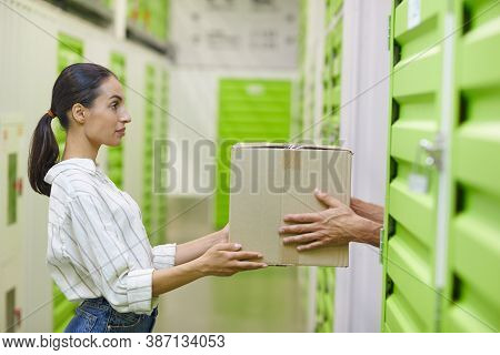 Side View At Young Woman Holding Cardboard Box And Handing In To Man While Packing Self Storage Unit