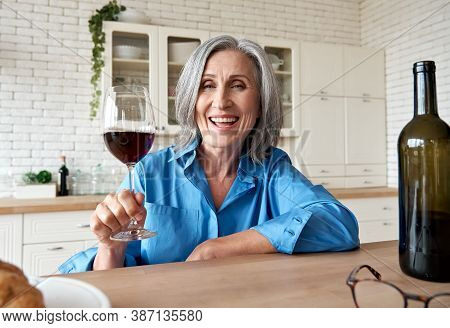 Happy 60s Mature Woman Drinking Wine Video Calling Friend At Home. Old Middle Aged Lady Holding Glas