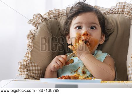Adorable Little Funny Girl Of 12 Months Eating Spaghetti With Spoon While Sitting In High-powered Ch