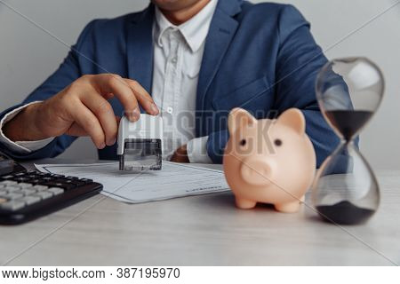 Businessman Approves Contract In Office. Pink Piggy Bank With Stack Of Coins And Hourglass On Desk. 