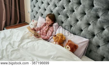 A Cute Small Happy Girl Lying With Teddybears In A Huge Bed Covered With Blanket Smiling And Reading
