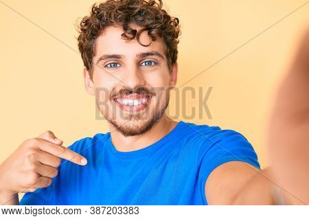 Young caucasian man with curly hair wearing casual clothes taking a selfie pointing finger to one self smiling happy and proud 