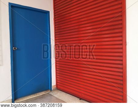 Two Doors Next To Each Other, Small Blue Wooden Door And Big Red Metal Door. Blue For People And Red