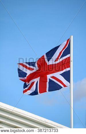 Union Jack Flag On Bright Blue Sky Background. Blown Away By Wind. United Kingdom Flag With Three Co