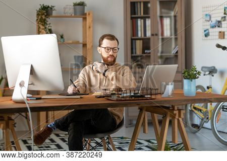 Young serious male freelance designer looking at laptop display while having drink and using tablet and stylus for drawing new logo