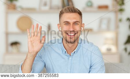 Greeting Concept. Portrait Of Smiling Handsome Man Waving Hello With Hand Looking At Camera, Sitting