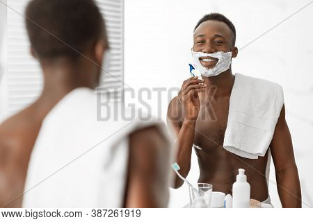 Shirtless African Guy Shaving Face Using Shave Foam And Razor Standing Near Mirror In Modern Bathroo