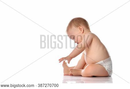 Cute Little Baby In Diapers Examines The Feet And Toes With Interest While Sitting On The Floor. Stu