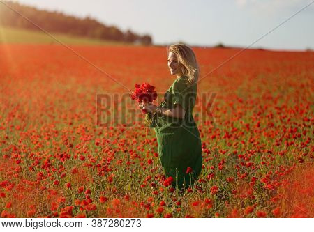 Beautiful Blonde Pregnant Woman In Long Green Dress With A Bunch Of Flowers In Hand, In The Middle O