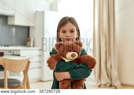 A Small Cute Girl Tightly Holds Her Fluffy Toy While Looking Into A Camera Stainding In A Spacy Kitc