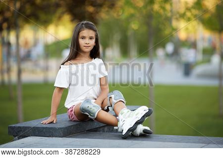 Happy Cute Little Child Girl In Roller Skates Loooking At The Camera In Summer Park.