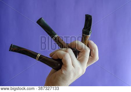 A Male Hand Holds Three Smoking Pipes On A Blue Background. Occasional Smoking Pipes Bend From Heath