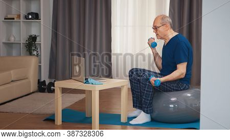 Senior Man Training In Living Room With Dumbbells During Online Fitness Program. Old Person Pensione