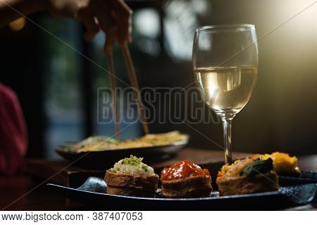 Close up shot of female hand enjoying Japanese cuisine various side dishes by chopstick, inari sushi set and white wine in restaurant with sun rays