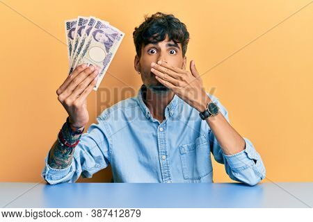Young hispanic man holding japanese yen banknotes sitting on the table covering mouth with hand, shocked and afraid for mistake. surprised expression 