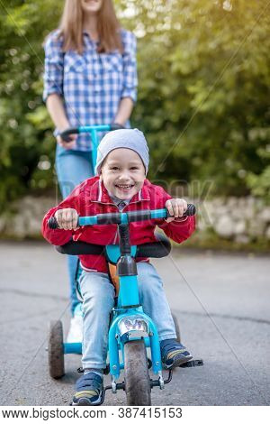 Young Mother Is Pushing A Childs Tricycle With A Toddler Boy On A Walk. Concept Of Learning To Ride 