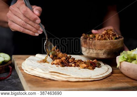 closeup of a young caucasian man preparing a durum or a burrito, with chicken meat cooked with different vegetables such as onion or red and green pepper, and fresh lettuce and raw cucumber