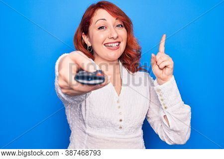 Young beautiful redhead woman changing television channel using tv remote control smiling with an idea or question pointing finger with happy face, number one