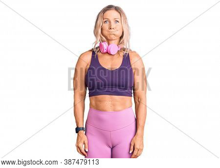 Middle age fit blonde woman wearing gym clothes and using headphones puffing cheeks with funny face. mouth inflated with air, crazy expression. 