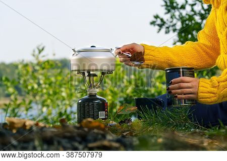 Young Beautiful Girl In A Yellow Sweater Makes Coffee In The Forest On A Gas Burner. Making Coffee O