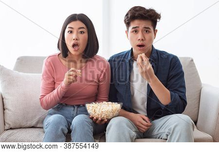 Shocked Asian Couple Watching Movie On Tv Eating Popcorn Sitting On Couch In Living Room At Home. Ja