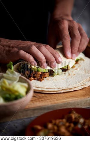 closeup of a young caucasian man preparing a durum or a burrito, with chicken meat cooked with different vegetables such as onion or red and green pepper, and fresh lettuce and raw cucumber