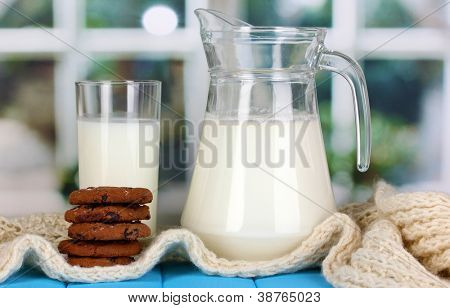 Jarra e copo de leite com biscoitos em malhas de gola alta na mesa de madeira no fundo da janela
