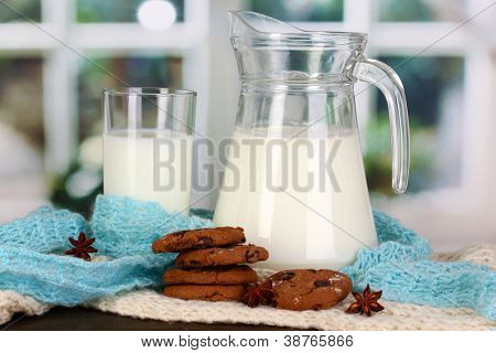 Jarra e copo de leite com biscoitos em malhas de gola alta na mesa de madeira no fundo da janela