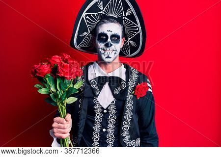 Young man wearing mexican day of the dead costume holding roses looking positive and happy standing and smiling with a confident smile showing teeth 