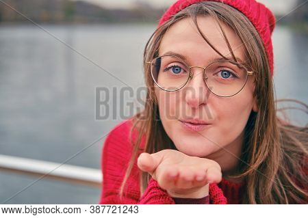 A Young Woman In A Red Sweater Sends An Air Kiss. Romance On The Shore Of An Winter Lake