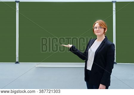 Woman Teacher Smiles And Points To An Empty Blackboard, Copy Space
