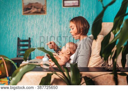 Young Mother Is Sitting On The Couch With The Baby, And Feeds It With A Spoon. There Is A Feeding Bo