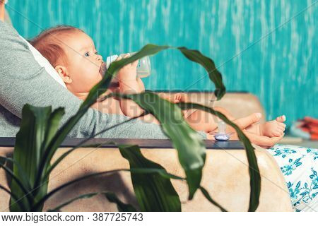 A Mother Is Sitting On The Couch With A Baby Who Is Eating Formula From A Bottle. Side View, From Ho