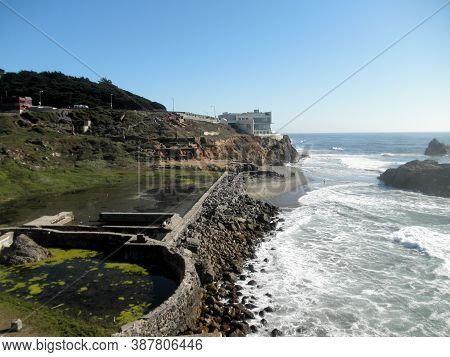Waves Roll Towards Shore Of Ocean Beach With Cliff House And The Ruins Of The Sutro Bath House In Fo