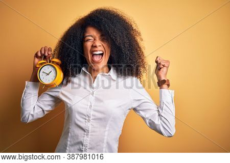 Young african american woman with afro hair holding classic alarm clock over yellow background screaming proud and celebrating victory and success very excited, cheering emotion