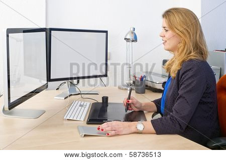Young woman sitting behind a large desk and a dual screen computer, using a graphic tablet and track pad during design work