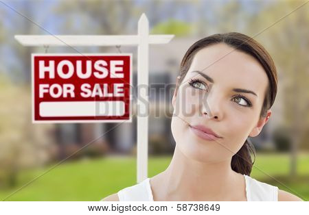 Thoughtful Pretty Mixed Race Woman In Front of Home and House For Sale Real Estate Sign Looking Up and to the Side.