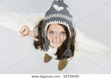 Pretty young woman in warm winter woollen pullover and grey cap, looking up and smiling while making victory sign, isolated on grey background.
