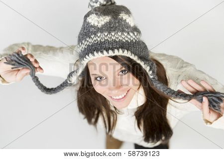 Beautiful young woman in white winter pullover and grey cap, looking up with raised arms and pretty smile, isolated on grey background.