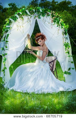 Beautiful bride with chaming red hair sitting under the wedding arch. Wedding dress and accessories. Wedding decoration. 
