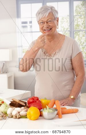 Happy grandmother standing in kitchen using fresh raw materials to prepare healthy food.