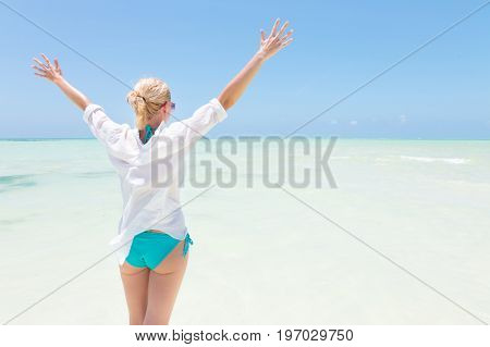 Happy woman, arms rised, enjoying, relaxing joyfully in summer at white tropical beach. Beautiful caucasian model wearing white beach tunic on vacations on Paje beach, Zanzibar, Tanzania.