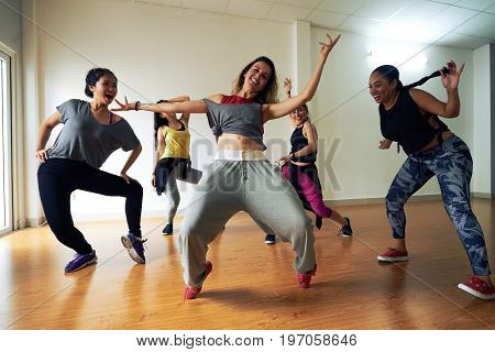 Full length portrait of pretty hip-hop dancer posing for photography with wide smile while standing on tiptoes, her colleagues dancing around her