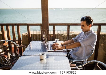 Thoughtful Young Asian Man Using Laptop In Cafe With View On Sea. Freelance Work