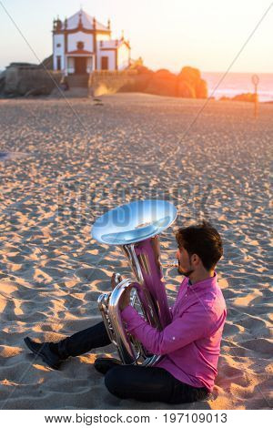 Young man playing the trumpet on the sea coast. Tuba instrument. In the background is the Chapel Senhor da Pedra at Miramar Beach, Atlantic ocean, Porto, Portugal.