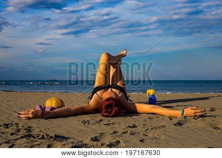 Young good looking tanned redhead laying on a sandy beach with arms resting and leg resting and happily posing with arms widespread with sun cream and hat on the side and blue sky and sea in the background during summer