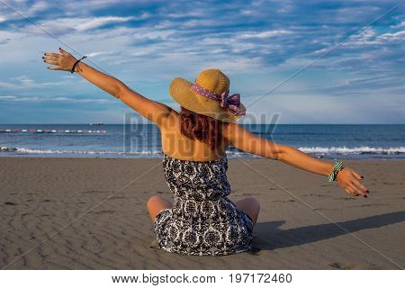 Beautiful girl woman in dress and hat sitting on the beach closeup with back turned and arms widespread with blue sea and sky in the background